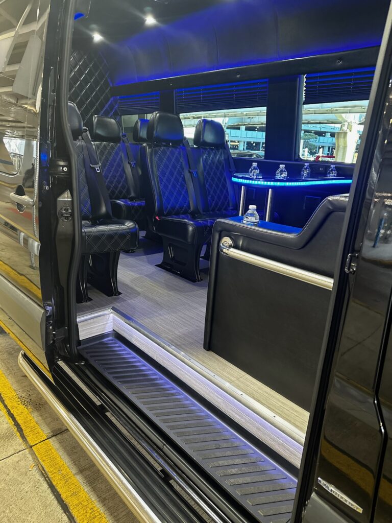 Open door of a luxury vehicle showing black leather seats, ambient blue lighting, and a mini bar with bottled water.