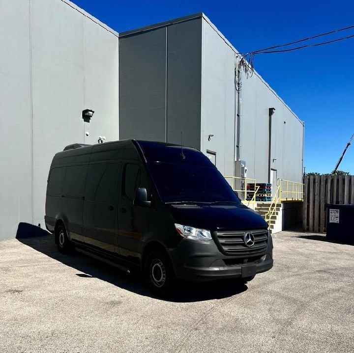 A black Mercedes-Benz van is parked in an industrial area beside gray buildings under a clear blue sky.