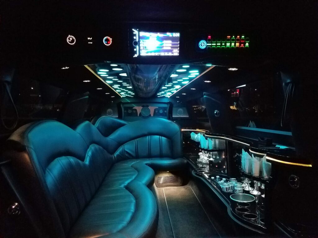 Interior of a luxury limousine showing plush blue seats, a bar area with glassware, ambient LED lighting on the ceiling, and a control panel above the seats.